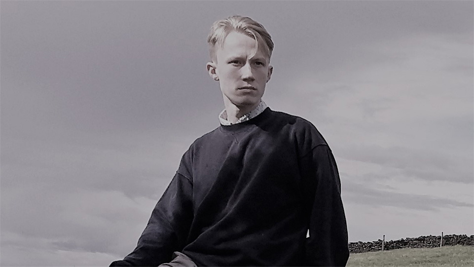 A man wearing a dark jumper, looking beyond the camera, sitting in the countryside.